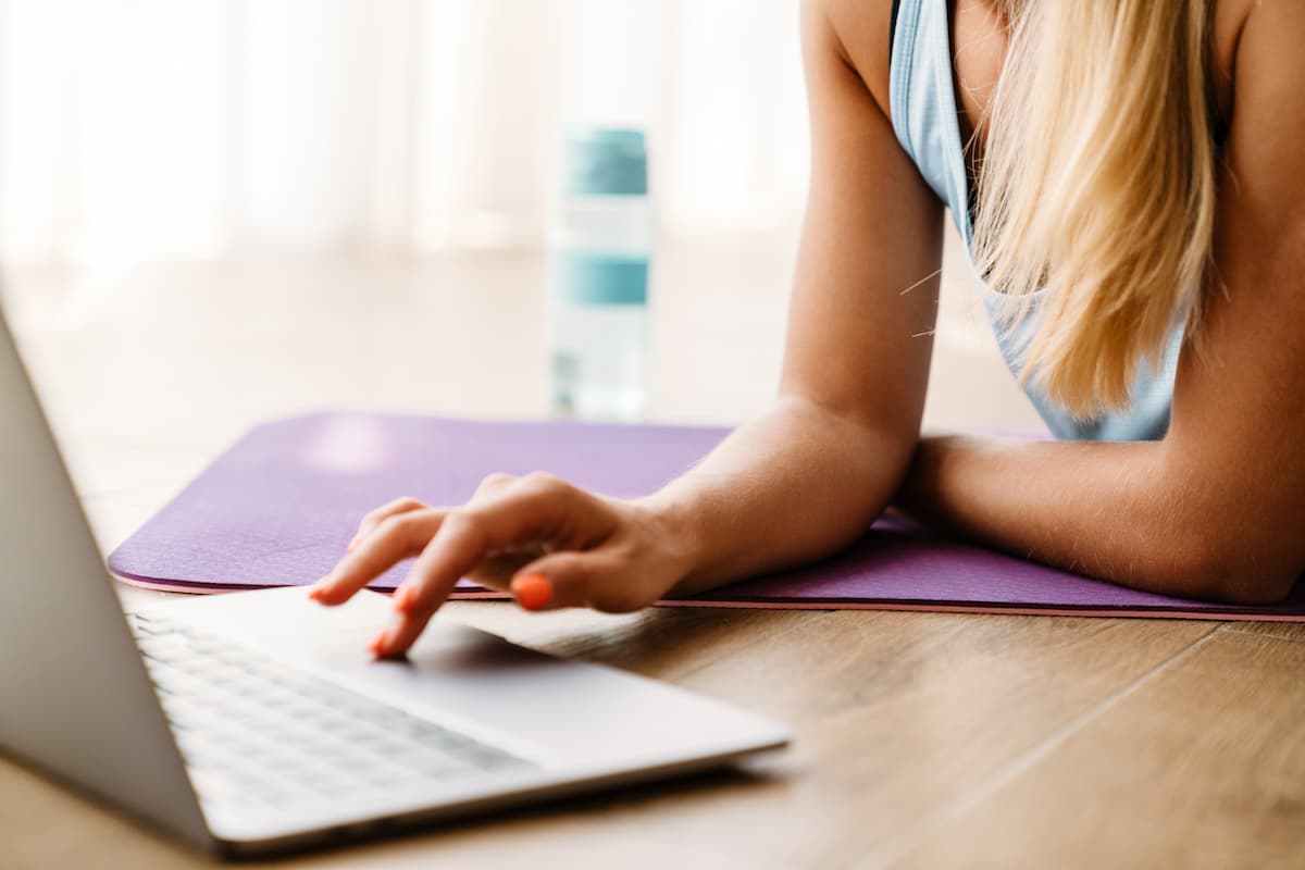 Woman with blonde hair lying on yoga mat looking at laptop